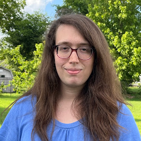 Mikayla, smiling outdoors in front of a row of trees